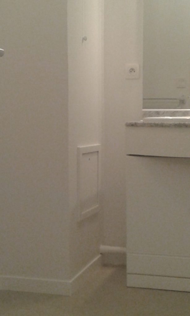 Partie de la salle de bain avec beaucoup d'angles (le mur fait un angle, il y a aussi un meuble avec un mirroir, un tuyau) peint en blanc, après application d'une toile de verre